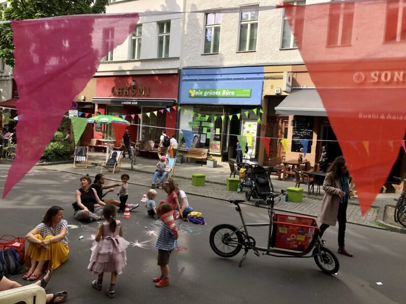 Auf dem Bild ist das Wahlkreisbüro abgebildet. Im Vordergrund sieht man die Wrangelstraße, die von Erwachsenen und Kindern bespielt wird. 
